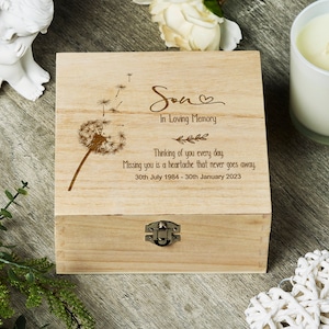 Puede incluir: Una caja de madera clara con cierre metálico. La caja está grabada con un diseño de diente de león y las palabras "Son, In Loving Memory". Texto adicional incluye "Thinking of you every day" y fechas.