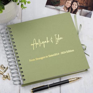 May include: A green spiral bound photo album with gold lettering that reads "Aaliyah & Isha From Strangers to Sister 2014 - 2024 Edition".