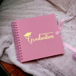 May include: Pink spiral bound scrapbook with a gold graduation cap and the text "Congratulations on your Graduation".