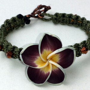 May include: A green crocheted bracelet with copper beads and a purple and white flower charm.