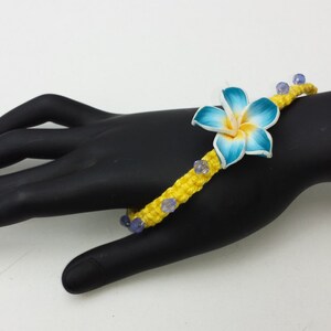 May include: A yellow macrame bracelet with blue and white flower accents and purple beads.