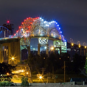 May include: A nighttime view of a bridge lit up in red, white, and blue lights. The bridge is a steel structure with a large arch in the middle. The bridge is over a body of water, and there are trees and buildings in the background.