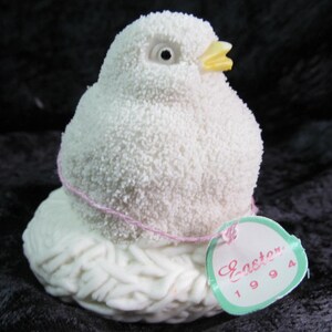 May include: White ceramic chick figurine with a yellow beak and a pink string around its neck. The chick is sitting in a white nest and has a tag that says "Easter 1994".