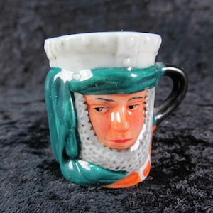 May include: A small, decorative ceramic mug featuring a painted face with orange skin, a white and green head covering, and a black handle. The mug has a white rim and a detailed, hand-painted design.