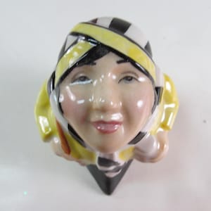May include: A ceramic figurine of a person with a smiling face, wearing a yellow and black striped head covering and a yellow jacket. The figurine has a black base and is set against a white background.