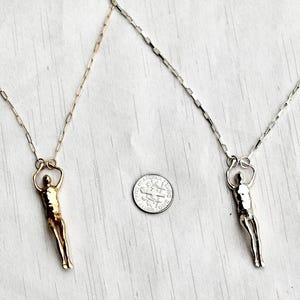 A gold-colored and a silver human sculpture pendant on thin chains are displayed next to a U.S. dime for scale.