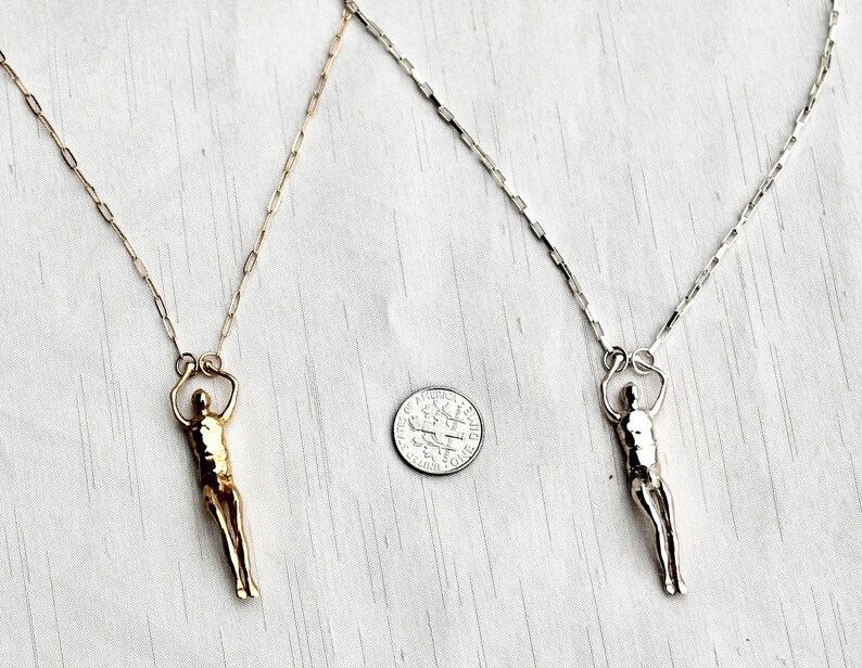 A gold-colored and a silver human sculpture pendant on thin chains are displayed next to a U.S. dime for scale.