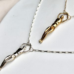 Two human sculpture pendants, one silver and one gold, hang from thin chains against a white background.
