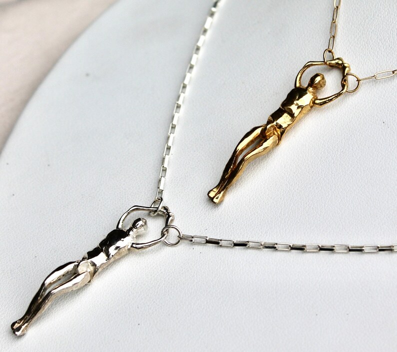 Two human sculpture pendants, one silver and one gold, hang from thin chains against a white background.