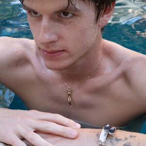 A man in a pool, with wet hair, wearing a gold alloy necklace with a small, stylized human figure pendant.