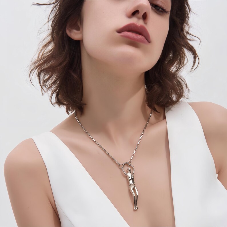 A woman wearing a silver-colored necklace with a stylized, reaching human figure pendant on a white top.