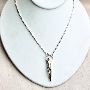 A silver human sculpture pendant on a thin chain is displayed on a white jewelry stand.