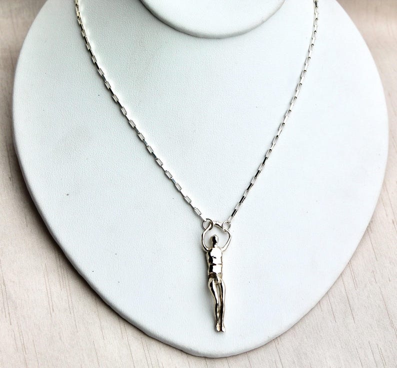 A silver human sculpture pendant on a thin chain is displayed on a white jewelry stand.