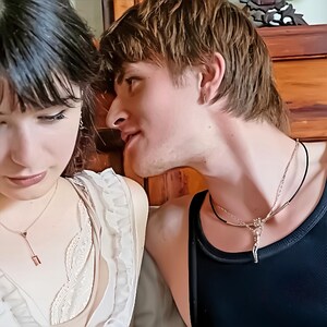 A man and woman, the man wearing a black cord necklace with a silver-colored reaching human figure pendant.