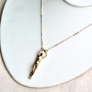 A gold-colored human sculpture pendant on a thin chain is displayed on a white jewelry stand.