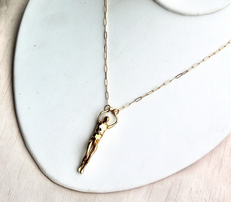 A gold-colored human sculpture pendant on a thin chain is displayed on a white jewelry stand.