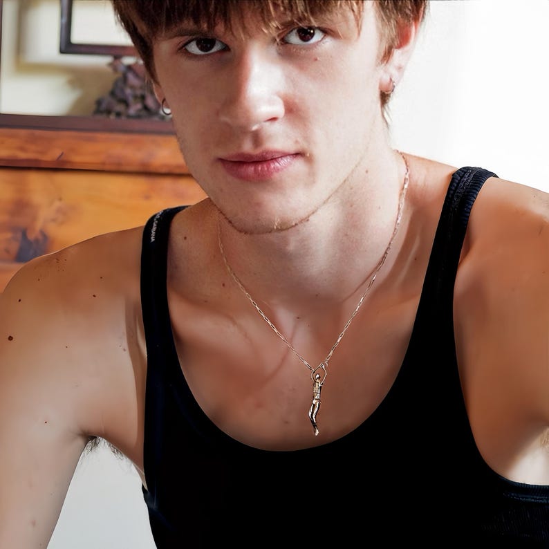 A person in a black tank top wears a gold-colored Reaching Man pendant on a thin chain, visible from the chest up.