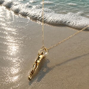 A gold-colored human sculpture pendant on a thin chain lies on wet sand with ocean waves in the background.