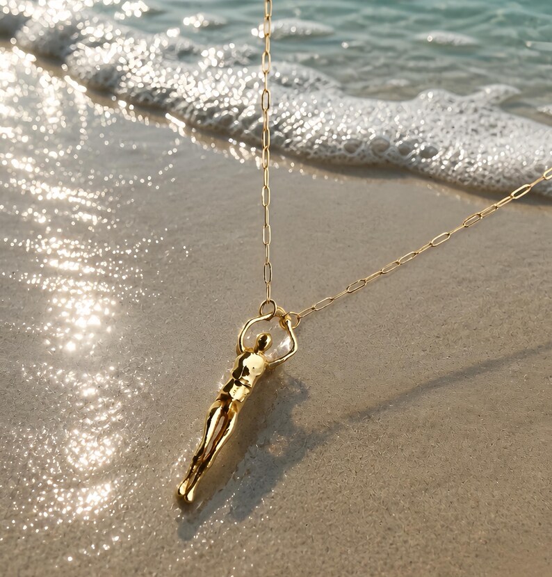 A gold-colored human sculpture pendant on a thin chain lies on wet sand with ocean waves in the background.