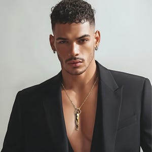 A man with short curly hair and a black blazer wears a gold-colored human sculpture pendant on a thin chain.