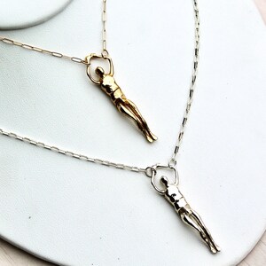 Two human sculpture pendants, one gold and one silver, hang from thin chains on a white surface.