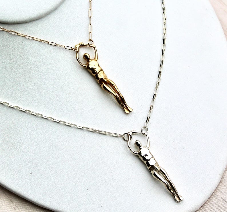 Two human sculpture pendants, one gold and one silver, hang from thin chains on a white surface.