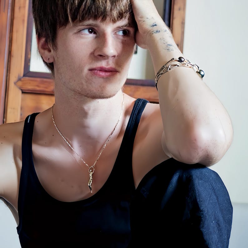 A person wearing a black tank top and a gold-colored Reaching Man pendant on a thin chain, with a silver bracelet on their right wrist.