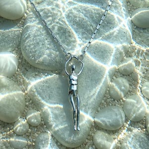 A silver human sculpture pendant on a thin chain is submerged in clear water with small white pebbles visible beneath.