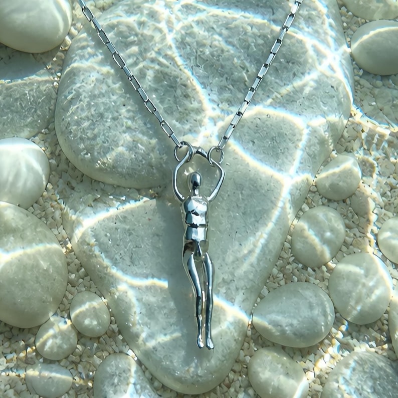 A silver human sculpture pendant on a thin chain is submerged in clear water with small white pebbles visible beneath.