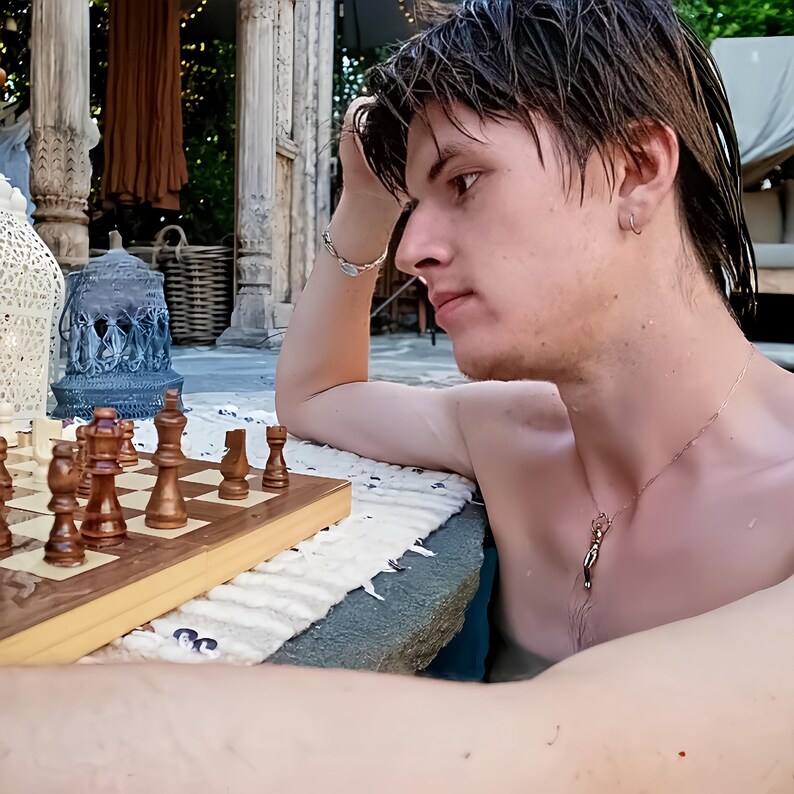 A man in a pool, with wet hair, wearing a gold alloy pendant of a reaching human figure, looking at a chessboard.