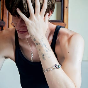 A person in a black tank top covers their face with one hand, wearing a silver Reaching Man pendant and a silver bracelet with dark beads.