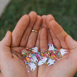 May include: A close-up of cupped hands holding a collection of confetti. The confetti includes white tooth shapes, gold coins, and colorful glitter in pink, blue, and green. A gold ring is visible on one finger.