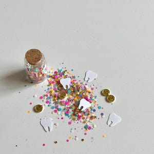 May include: A small glass bottle with a cork stopper filled with colorful confetti. Scattered around are white paper teeth and small gold coins on a white surface. The confetti includes pink, yellow, blue, and green specks.