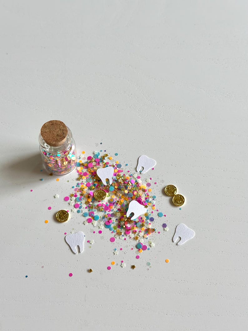 May include: A small glass bottle with a cork stopper filled with colorful confetti. Scattered around are white paper teeth and small gold coins on a white surface. The confetti includes pink, yellow, blue, and green specks.