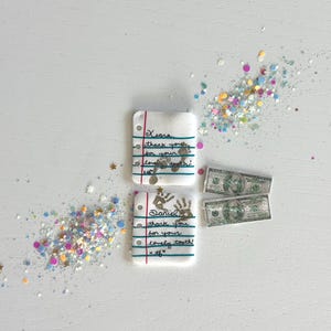 May include: Two miniature notebooks with handwritten notes, alongside two toy dollar bills and scattered glitter. The notebooks read "Kiara, thank you for your lovely tooth!" and "Daniel, thank you for your lovely tooth!"