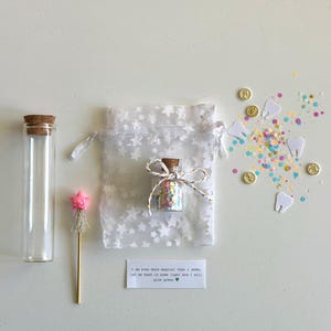 May include: A collection of whimsical items, including a small glass bottle filled with colorful confetti, a clear glass tube with a cork stopper, a star-topped wand, and a white bag with star patterns. Scattered around are gold coins, paper teeth, and confetti. A small card reads, "I am even more magical than I seem, let me bask in some light and I will glow green."