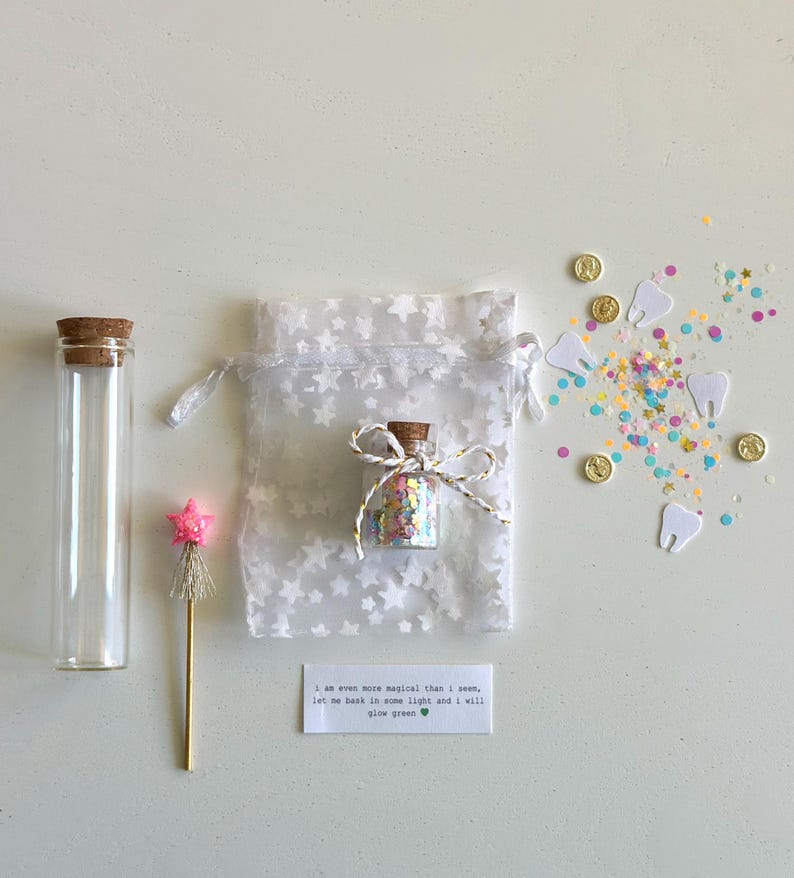 May include: A collection of whimsical items, including a small glass bottle filled with colorful confetti, a clear glass tube with a cork stopper, a star-topped wand, and a white bag with star patterns. Scattered around are gold coins, paper teeth, and confetti. A small card reads, "I am even more magical than I seem, let me bask in some light and I will glow green."