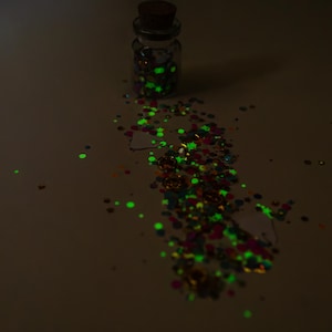 May include: A small glass bottle with a cork stopper, filled with colorful glitter. Some glitter is spilled around the bottle. The glitter glows bright green in the dark, with other colors visible.