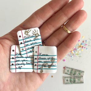 May include: A hand holding three small, white, notepad-shaped keepsakes with handwritten messages and gold-colored handprints and footprints. The notes say "thank you for your lovely tooth!" and are decorated with red and blue lines.