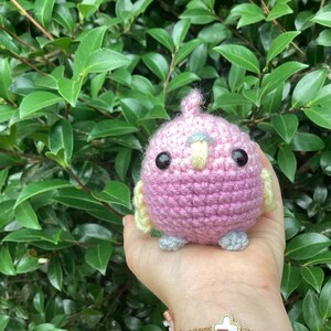 May include: A pink crocheted bird with yellow and gray accents. The bird has black eyes and a yellow beak. The bird is being held in a person's hand.