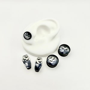 May include: A set of four black and white bat themed ear gauges. Two gauges are round and two are coffin shaped. The gauges are made of clear resin with black bats embedded in the resin. The gauges are displayed on a white background.