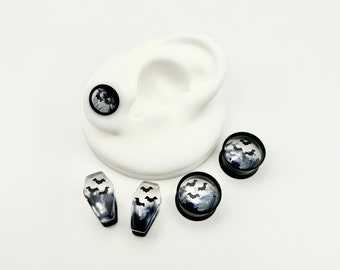May include: A set of four black and white bat themed ear gauges. Two gauges are round and two are coffin shaped. The gauges are made of clear resin with black bats embedded in the resin. The gauges are displayed on a white background.