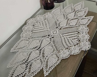 Könnte beinhalten: Ein zartes, gehäkeltes Tischläufer in hellgrau. Der Läufer hat ein florales Design mit filigranen Details.