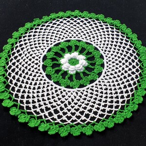 Puede incluir: Un tapete circular hecho a mano con un diseño calado blanco, un borde y acentos verdes. El centro tiene una flor blanca rodeada de hojas verdes. Esta pieza decorativa es perfecta para añadir un toque de encanto vintage a cualquier hogar.