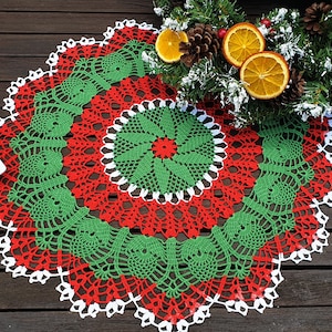 Puede incluir: Un encaje de ganchillo redondo de color verde, rojo y blanco con un diseño circular. El encaje está sobre una superficie de madera.