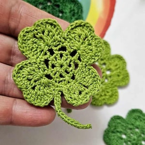 May include: Close-up of several handmade green crochet four-leaf clovers. The clovers are detailed with visible stitching and a textured appearance. A rainbow graphic is visible in the background. These clovers are ideal for St. Patrick's Day or other craft projects.