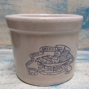 May include: A white ceramic crock with a blue design featuring a teddy bear holding an American flag. The design also includes the words "America", "Peace", "Freedom", "Liberty", "U.S.A.", and "Life".