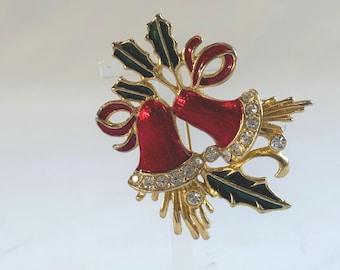May include: A Christmas-themed brooch featuring two red bells with rhinestone accents, gold-tone metal, and green holly leaves. The bells have red bows and are connected by gold stems. A festive holiday accessory.