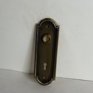 May include: A brass door plate with a keyhole and a round hole for a doorknob. The plate has a decorative border and is in a vintage style.