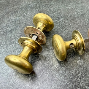 May include: Four antique brass door knobs with a classic design. Each knob has a rounded shape and a circular backplate. The brass has a warm, golden hue, and the knobs are arranged on a textured gray surface.